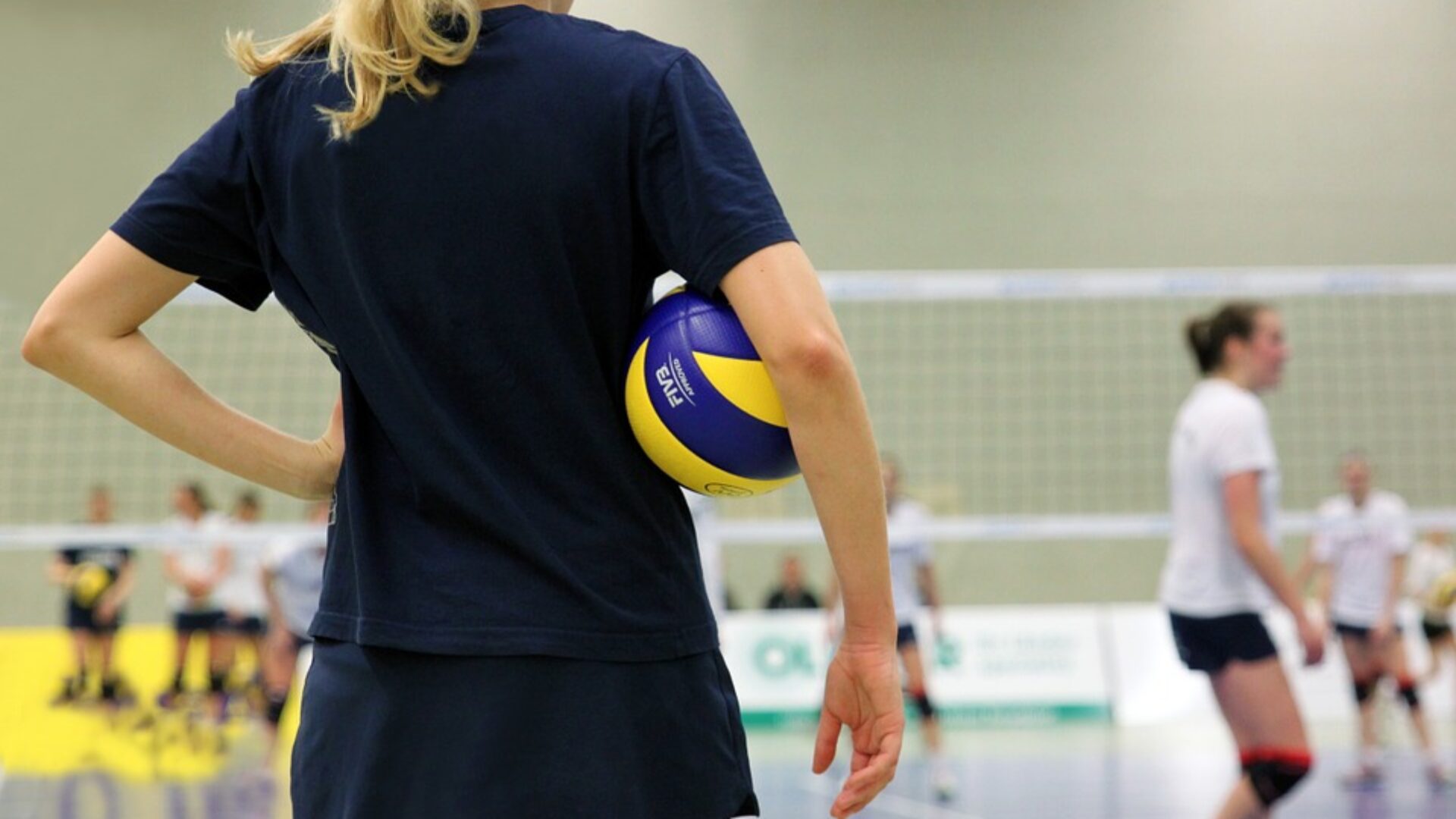 Vrouw op volleybalveld met een volleybal onder haar arm.