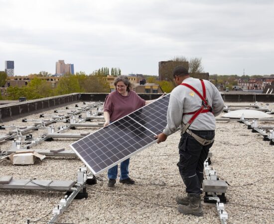 Musetta Blaauw (initiatiefnemer coöperatie GOED) legt samen met de installateur de eerste zonnepaneel op het dak van Lentis aan de Hereweg neer.