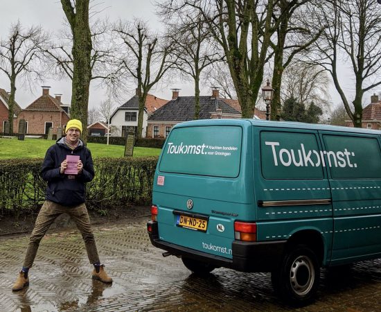 Toukomstcollega Thijs staat vrolijk bij de Toukomstbus met een Toukomstmuts op zijn hoofd.