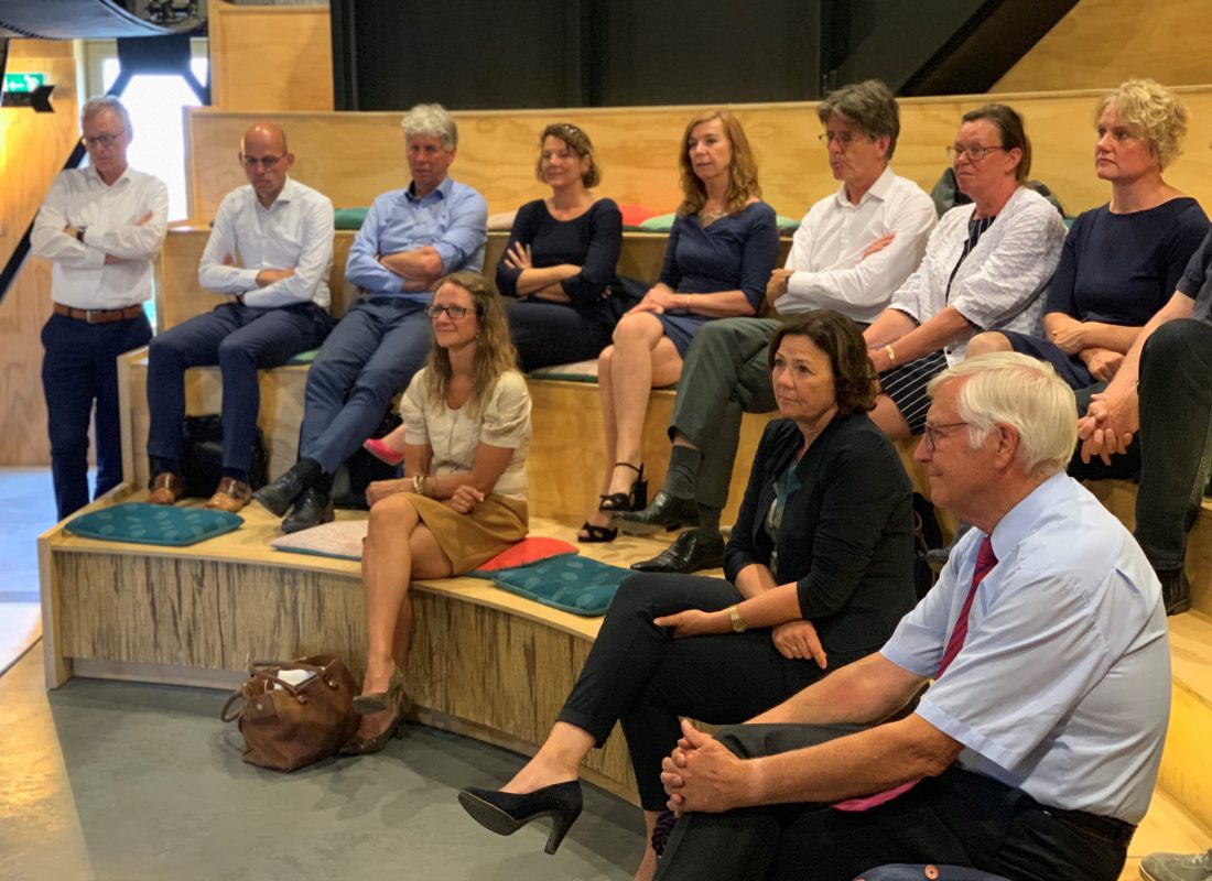 Elf stakeholders, waaronder directeur Siem Jansen, zitten op een tribune aandachtig te luisteren naar iemand die iets presenteert.