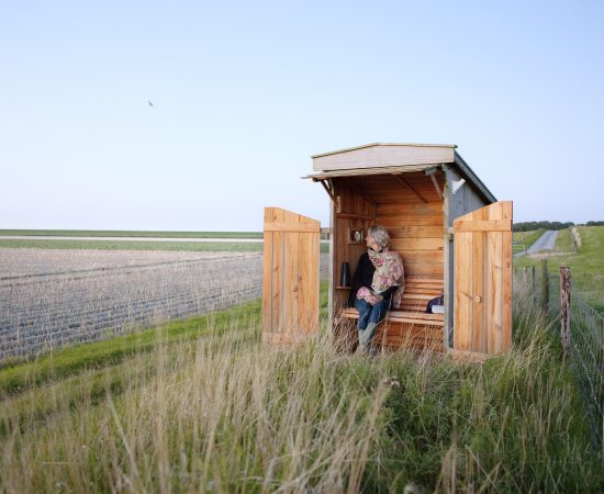 Vrouw in een klein houten hutje tussen lang uitgestrekte akkers.