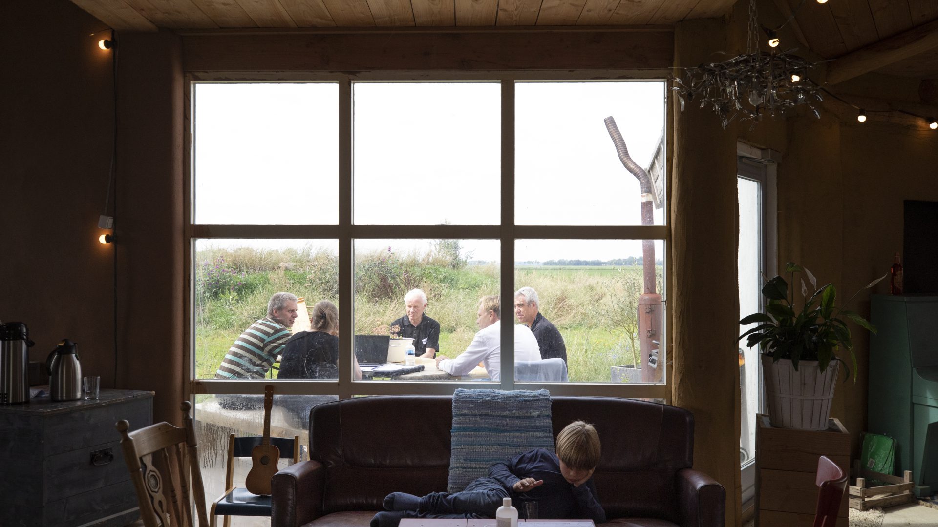 Binnenhuis ligt een kind op bank met op de achtergrond door het raam zichtbare buitenoverleg met vijf personen