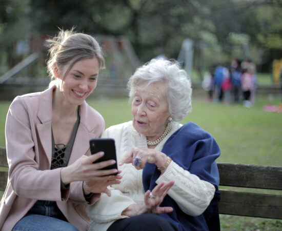 Decoratieve foto van een bejaarde vrouw en een vrouw van middelbare leeftijd die samen op een bankje naar een telefoon kijken